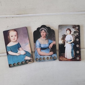 Op de afbeelding: Drie houten bladwijzers met portretten van historische figuren. De portretten zijn in blauw, wit en bruin. Elke bladwijzer heeft een reeks cirkelvormige gaten langs de onderrand.