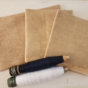 May include: Three beige fabric squares with a blue and white spool of thread and a white spool of thread on top.