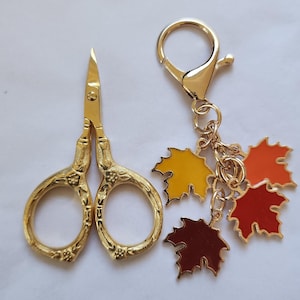May include: Gold-coloured embroidery scissors and a gold keychain with four autumn-themed maple leaf charms in yellow, orange, and red. The scissors have a floral design.