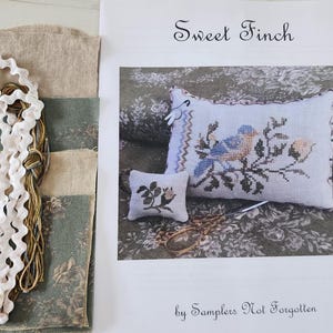 May include: A cross stitch pattern kit for a pillow featuring a bluebird perched on a branch with flowers. The kit includes fabric, floss, and instructions. The pattern is titled "Sweet Finch" by Samplers Not Forgotten.