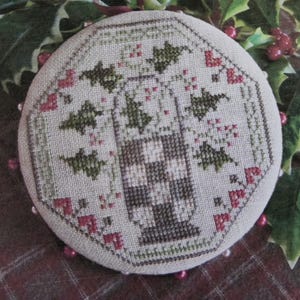 May include: A round, beige fabric ornament featuring a cross-stitch design of a central checkered urn surrounded by green holly leaves and small red berries. The circular piece is edged with a series of small, light-colored beads along the perimeter.