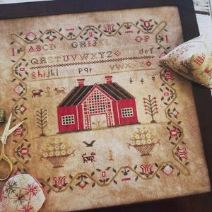 Puede incluir: Un sampler de punto de cruz enmarcado con una casa roja con tejado negro, rodeada de patrones florales y geométricos. El sampler incluye el alfabeto y elementos decorativos, con un pequeño alfiletero y tijeras.