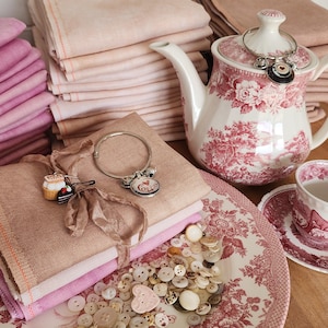 May include: A still-life arrangement featuring stacks of pink fabric, a floral-patterned teapot, and a matching teacup and saucer. A plate with a floral design holds a collection of buttons. A bracelet and charms are also present.