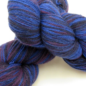 Puede incluir: Un ovillo de hilo en una combinación de colores azul y morado. El hilo es una mezcla de lana y seda y tiene una textura suave y peluda.
