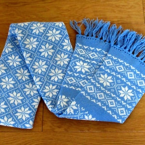 Peut inclure: Écharpe tricotée bleu clair et blanche avec un motif de flocons de neige. L'écharpe présente un motif répété de flocons de neige et de losanges blancs sur un fond bleu clair. Les extrémités de l'écharpe ont une frange.