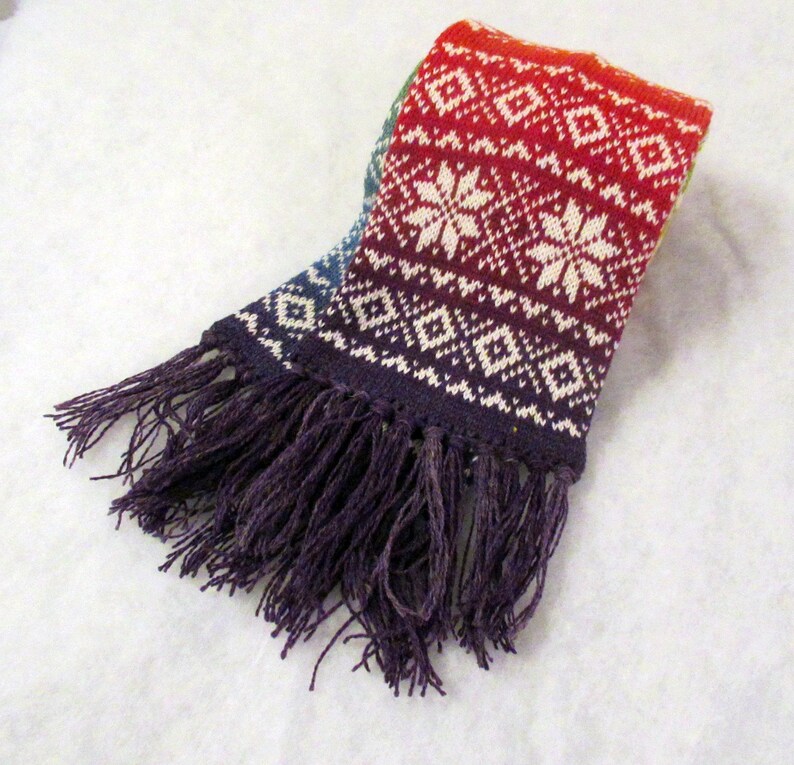 May include: A rainbow knit scarf with a white snowflake pattern and purple fringe.
