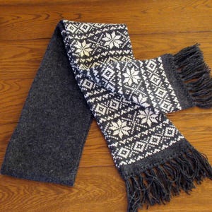 May include: A gray and white patterned scarf with a fringe on both ends. The scarf is made of a soft, knitted fabric.