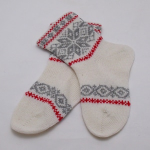 May include: A pair of off-white knitted socks with a snowflake pattern in gray and red. The socks have a red trim at the top and around the patterned sections. The design is a classic winter style.