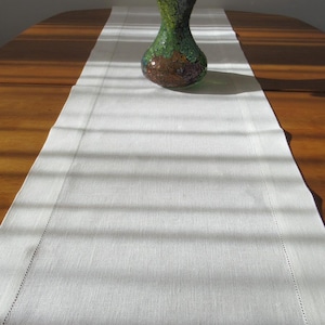 Può includere: Un runner da tavolo bianco con una texture sottile si trova su un tavolo di legno. Un vaso pieno di tulipani gialli si trova al centro del runner. Il vaso è fatto di vetro con un motivo a mosaico in tonalità di verde, marrone e oro.