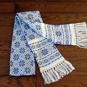 May include: A white and blue knitted scarf with a geometric pattern and white fringe on the ends.