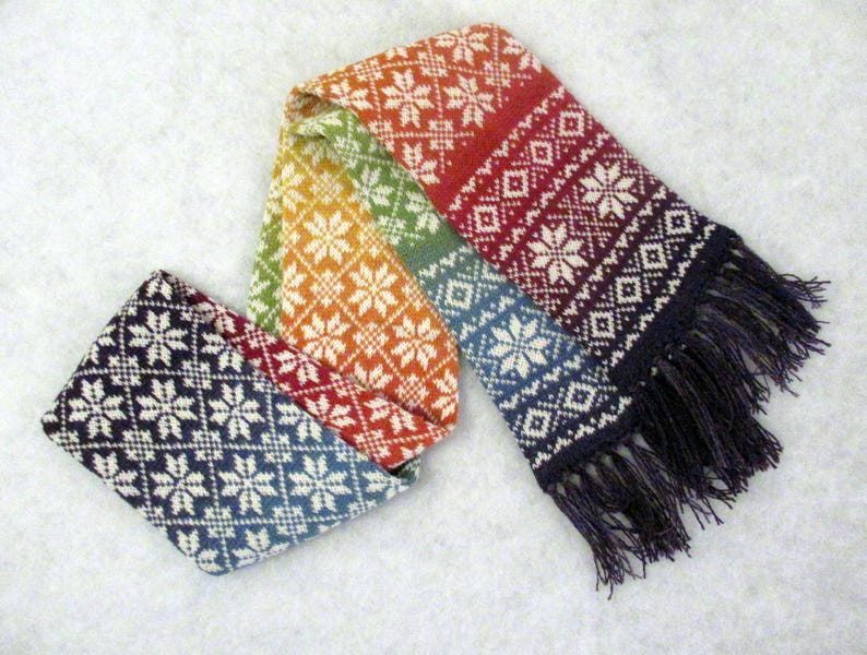 May include: A colorful knitted scarf with a geometric pattern and fringe. The scarf is made of wool and has a rainbow color scheme.