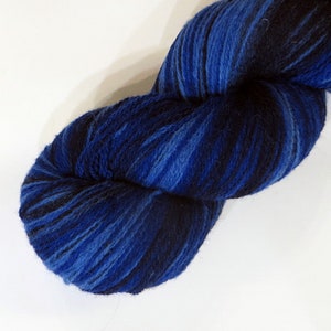 Puede incluir: Un ovillo de lana en tonos azules y negros. La lana parece suave y texturizada, con un efecto degradado de azul oscuro a claro. Ideal para proyectos de tejido o ganchillo.
