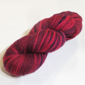 Puede incluir: Un ovillo de hilo en una combinación de colores rojo oscuro y morado oscuro. El hilo es una mezcla de lana y seda, y es suave y lujoso.