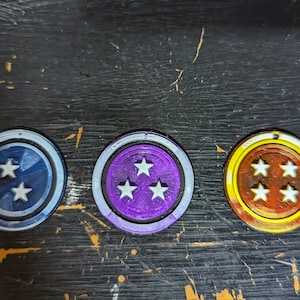 May include: Three round tokens with star designs. The tokens have a white border and a central design with white stars. The first token is navy blue with two stars, the second is purple with three stars, and the third is brown with four stars.