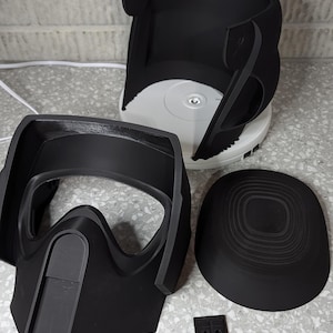 Può includere: Un set di parti per casco stampate in 3D di colore nero, tra cui una maschera per il viso, una cupola e un pezzo circolare. Le parti sono su una superficie grigia chiaro macchiata.