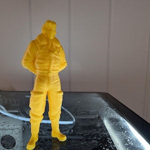 May include: A yellow 3D-printed figurine of a person in a flight suit, standing with hands behind the back. The figure has a detailed jacket, trousers, and boots. The figurine is on a reflective surface.