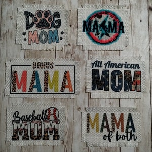 May include: A collection of burlap patches with various designs and text. The patches feature phrases like "Dog Mom," "Mama," "Bonus Mama," "All American Mom," "Baseball Mom," and "Mama of both." The text is in various colors and fonts, with some designs incorporating animal print and baseball imagery.