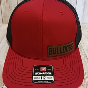 May include: Red and black trucker hat with a brown leather patch that says "BULLDOGS". The hat has a black mesh back and a red front with a black stitched brim. The hat also has a black label with white text that says "EST. 1970 RICHARDSON. ORIGINAL 112 TRUCKER ADJUSTABLE".