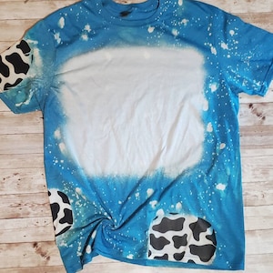 May include: A blue t-shirt with a white bleach design and black and white cow print patches on the sleeves and bottom.