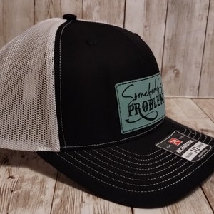 May include: Black and white trucker hat with a green patch that says "Somebody's Problem" in a cursive font.