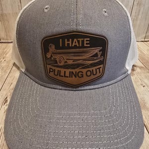 May include: Gray trucker hat with a white mesh back and a curved brim. The front features a brown leather patch with the words "I HATE PULLING OUT" above an illustration of a boat on a trailer.
