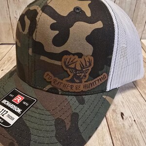 May include: Camouflage trucker hat with a brown leather patch featuring a deer head and the text "I'd Rather Be Hunting." The hat has a white mesh back and a curved brim. The Richardson brand label is visible.