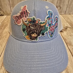 May include: A light blue trucker hat with a white mesh back. The hat features patches of a butterfly with the name "Dolly", a Highland cow, and a cactus. The hat has a curved brim and a snapback closure.