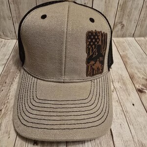 May include: A beige and black trucker hat with a brown leather patch featuring a silhouette of a person saluting in front of an American flag.