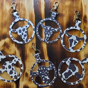 May include: Six cow print keychains with a cow head cutout. The keychains are made of leather and have a silver metal ring.