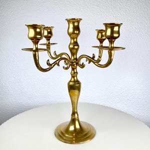 May include: A vintage brass candelabra with five candle holders. The candelabra has a central stem and curved arms, each holding a candle cup. The base is round and tiered. The overall height is approximately 10 inches.