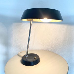 Op de afbeelding: Een zwarte bureaulamp met een zilveren steel en een ronde zwarte voet. De lamp heeft een gebogen, zwarte lampenkap met een gouden binnenkant. De lamp is ingeschakeld en verlicht de gouden binnenkant. De lamp staat op een lichtgekleurde, ronde tafel.