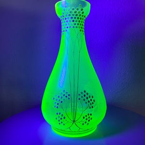 Op de afbeelding: Een gloeiende groene glazen vaas met een uitlopende voet en een smalle hals. De vaas heeft een decoratief patroon van zwarte lijnen en cirkels. De vaas is ongeveer 25 cm hoog.