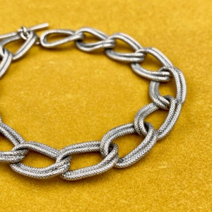 Op de afbeelding: Zilverkleurige schakelarmband met een getextureerd oppervlak. De armband heeft grote, ovaalvormige schakels die zijn verbonden door een tuimelsluiting. De sieraden worden op een gele achtergrond gepresenteerd.
