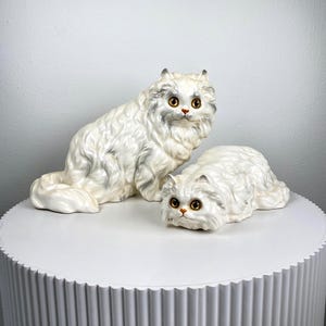 Può includere: Due figurine in ceramica bianca di gatti persiani. I gatti sono seduti e sdraiati. Il gatto seduto ha gli occhi dorati e il gatto sdraiato ha gli occhi marroni.
