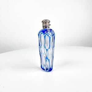 May include: A small, vintage perfume bottle with a silver-colored cap. The bottle is made of clear glass with a striking blue geometric design. The bottle's shape is elongated and cylindrical, perfect for display.