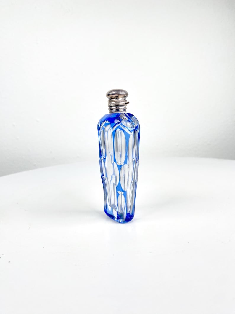 May include: A small, vintage perfume bottle with a silver-colored cap. The bottle is made of clear glass with a striking blue geometric design. The bottle's shape is elongated and cylindrical, perfect for display.