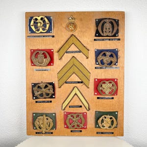 May include: A wooden display board showcasing military insignia and rank badges. The board features various badges with gold and bronze emblems, chevrons, and text labels. The badges are mounted on fabric squares. The background is white, and the board is light brown.