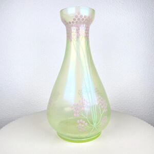 May include: An iridescent green glass vase with a flared top and a pear shape. The vase has a floral design in pink and green, with small pink circles and thin green stems. The vase is approximately 20 cm tall.
