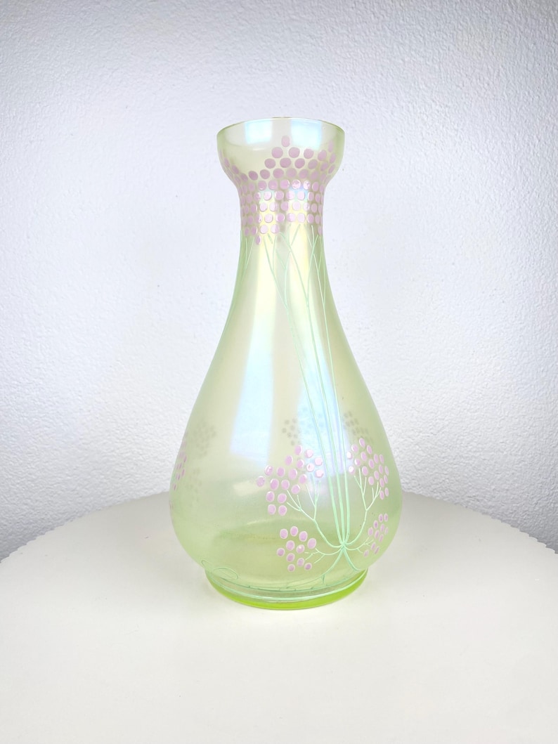 May include: An iridescent green glass vase with a flared top and a pear shape. The vase has a floral design in pink and green, with small pink circles and thin green stems. The vase is approximately 20 cm tall.
