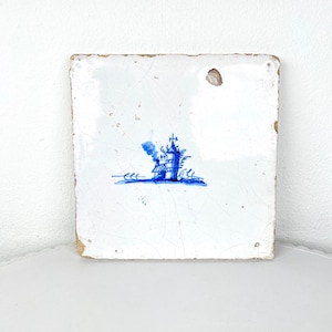 May include: Square, white ceramic tile featuring a blue illustration of a ship. The ship design includes a mast and smoke. The tile shows signs of age, with cracks and imperfections adding to its character.