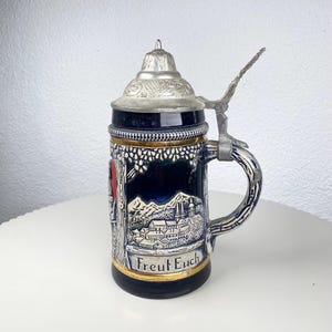 May include: A blue and white ceramic beer stein with a silver lid and handle. The stein features a detailed landscape scene and the text "Freut Euch". The rim is trimmed with gold.