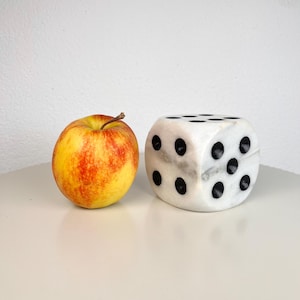 May include: A round apple with yellow and red skin next to a white marble die with black circular pips. The die features rounded corners and a smooth, polished stone surface, showing the top face with six pips and the side face with five pips.