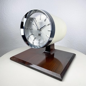 Könnte beinhalten: Eine Vintage Citizen Quartz Crystron Tischuhr. Die Uhr hat ein silbernes Zifferblatt mit schwarzen Zeigern und eine Chromlünette. Das Gehäuse der Uhr ist cremefarben und steht auf einem dunkelbraunen, rechteckigen Holzsockel.