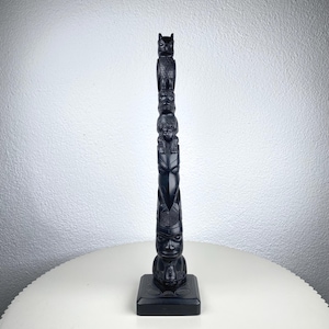 Op de afbeelding: Een zwarte totempaal met gebeeldhouwde figuren van een uil, menselijke gezichten en andere symbolische ontwerpen. De totempaal staat op een vierkante basis en wordt tentoongesteld op een witte tafel.