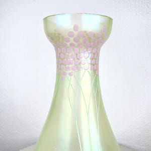 May include: A pearlescent green glass vase with a flared base and a narrow neck. The neck is decorated with pink polka dots and delicate green lines. The vase has a soft, iridescent sheen. A decorative item.