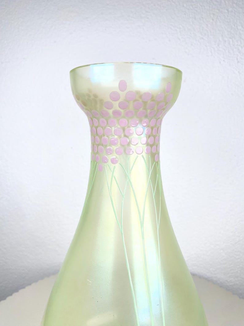 May include: A pearlescent green glass vase with a flared base and a narrow neck. The neck is decorated with pink polka dots and delicate green lines. The vase has a soft, iridescent sheen. A decorative item.