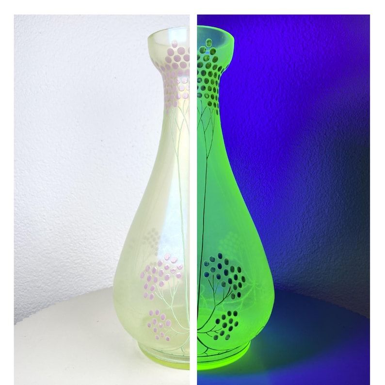 May include: A tall glass vase with a narrow neck and a rounded base. The vase is split into two sections: one pale yellow with pink floral patterns, the other glowing green with black tree and dot designs. It rests on a white surface.
