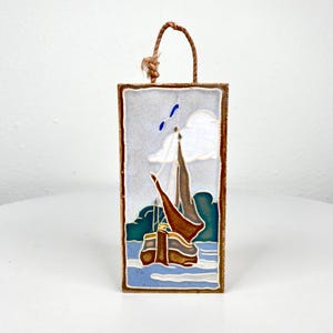 May include: A rectangular wooden plaque with a painted image of a sailboat on the water. The sailboat has a brown sail and a blue mast. The water is blue and there is a white cloud in the sky.