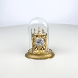 May include: A miniature gold-colored clock encased in a clear glass dome. The clock has a detailed, ornate design with a silver clock face. The base of the dome is also gold, creating a vintage, decorative piece.