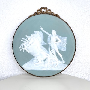 Puede incluir: Una obra de arte mural redonda y enmarcada con un fondo azul claro. La obra presenta un relieve blanco de una figura en un carro tirado por caballos. El marco es de metal oscuro y ornamentado.